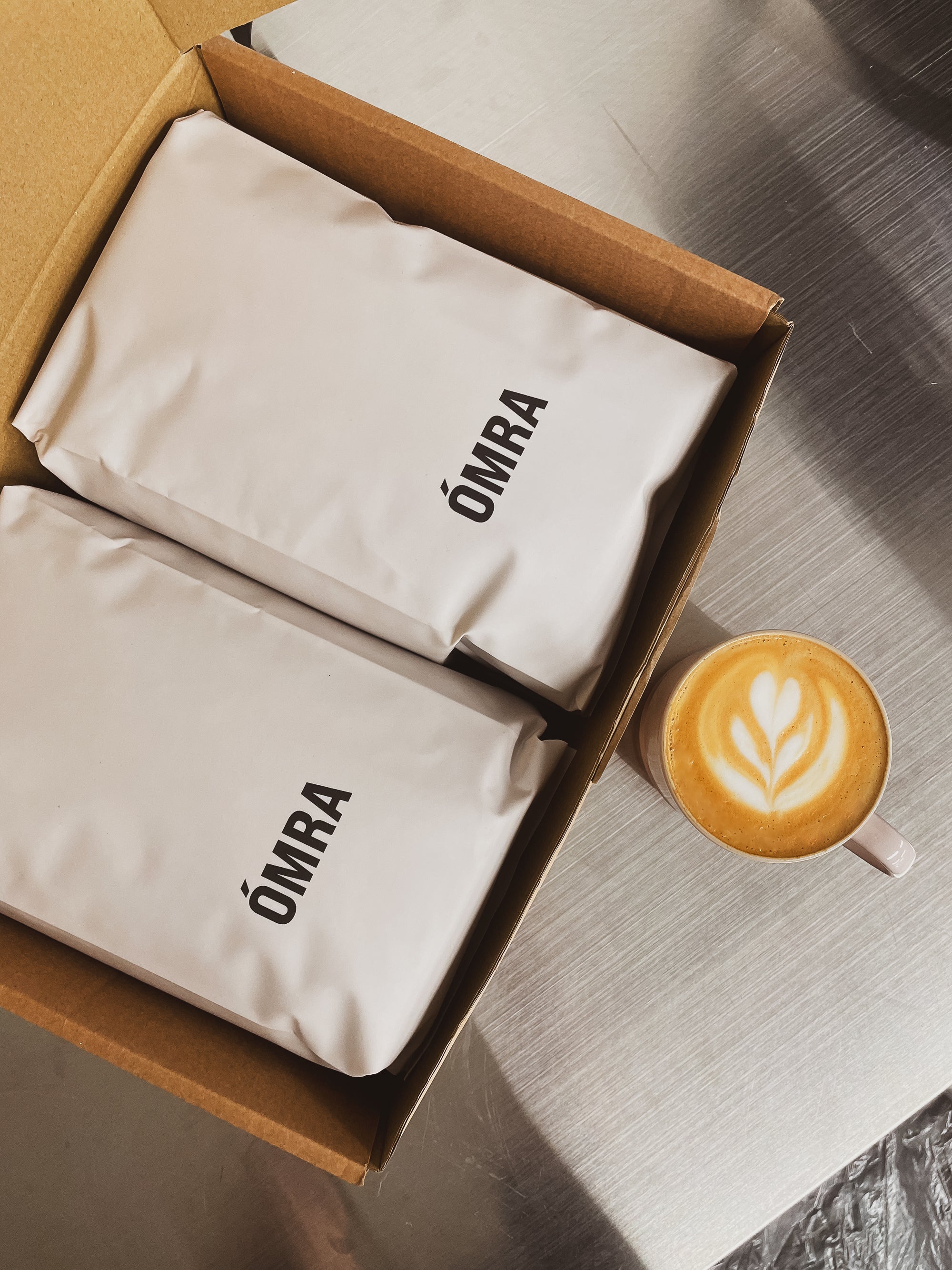 Two bags of freshly roasted Ómra specialty coffee on a table beside a freshly brewed flat white with latte art.